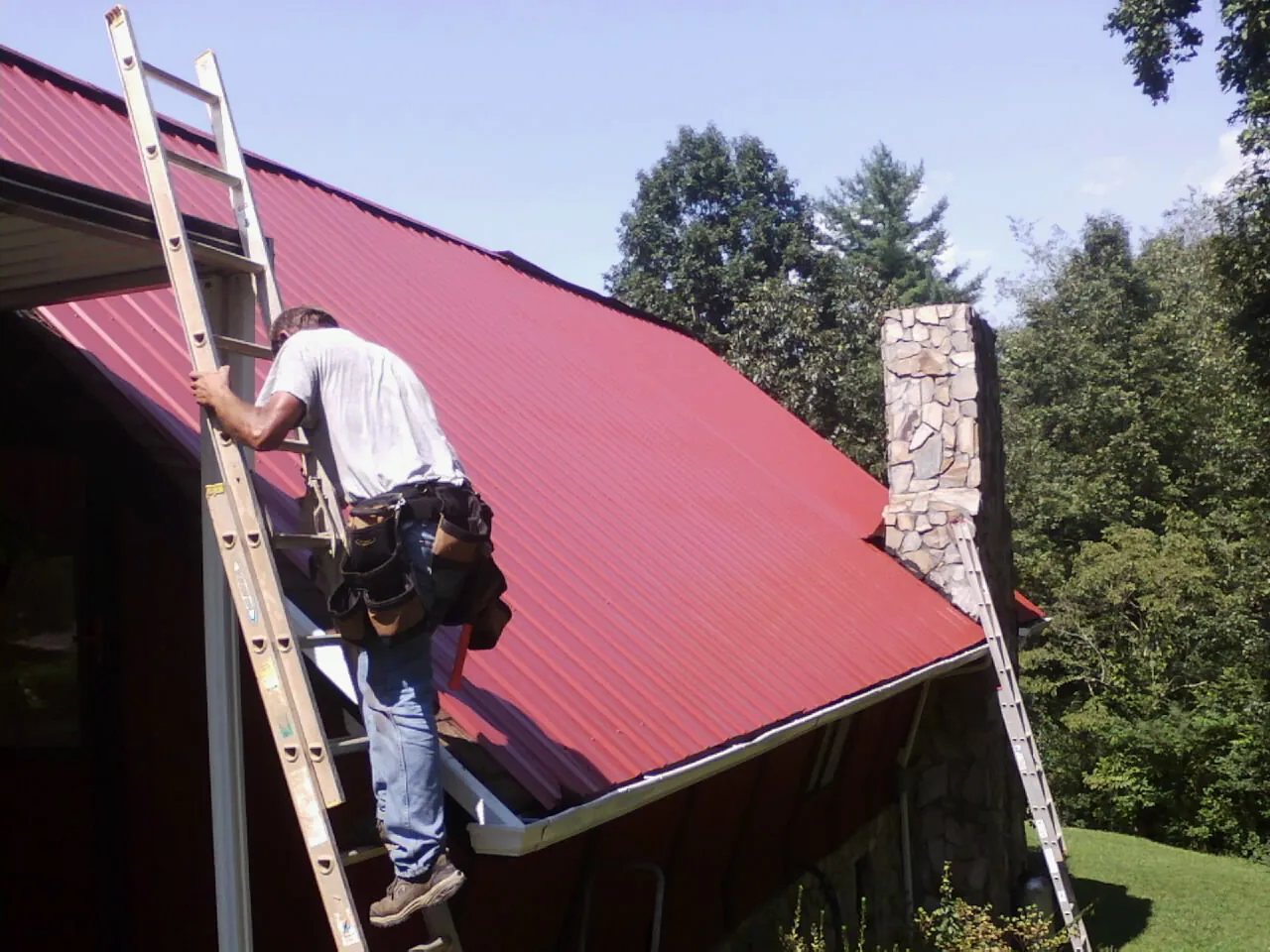 Roofer on a ladder beside a red metal roof and stone chimney — Soffit & Fascia Repair by Roofing Brothers in Gardendale